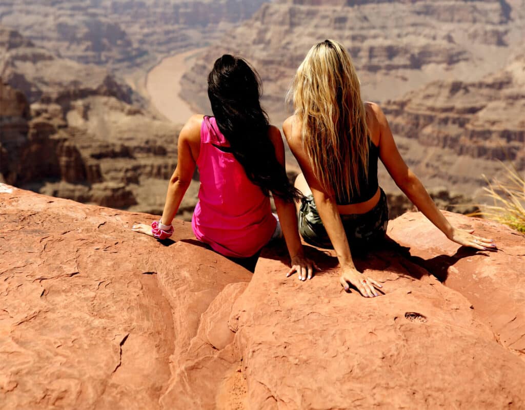 c0_hero_lg-1-2 Two women sit on a rocky cliff overlooking a vast canyon with a winding river.