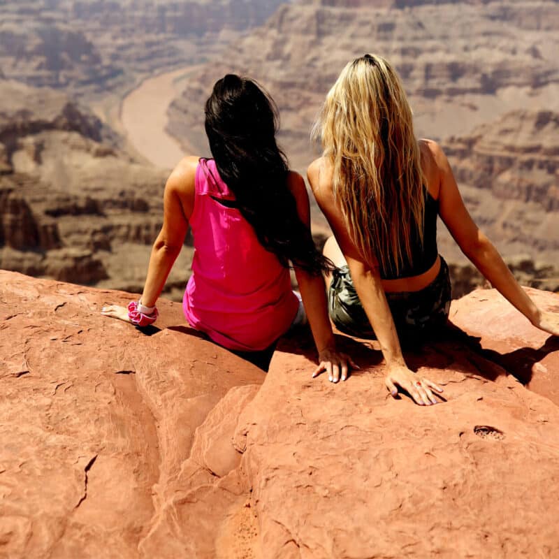 Two women sit on a rocky cliff overlooking a vast canyon with a winding river.