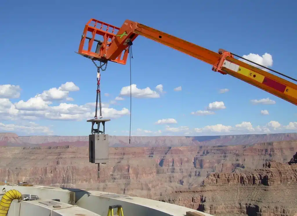 Grue orange soulevant une lourde pièce d'acier pendant la construction du Grand Canyon Skywalk, avec en arrière-plan les falaises pittoresques du canyon et le ciel bleu.