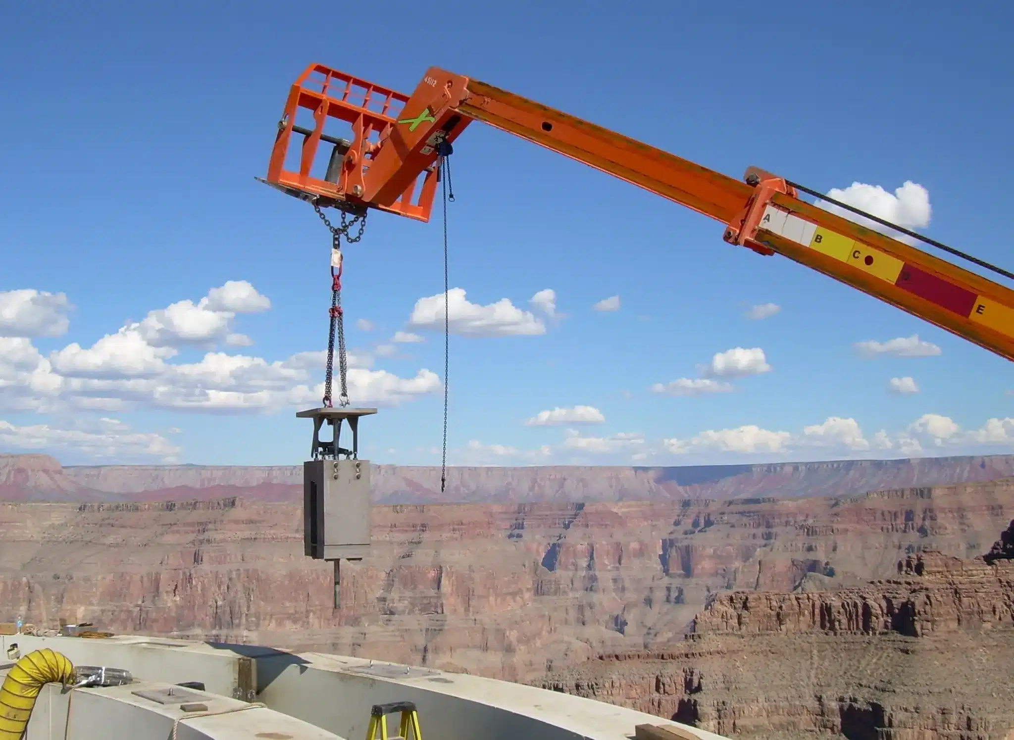Orange crane lifting a heavy steel component during construction of the Grand Canyon Skywalk, with scenic canyon cliffs and blue sky in the background.