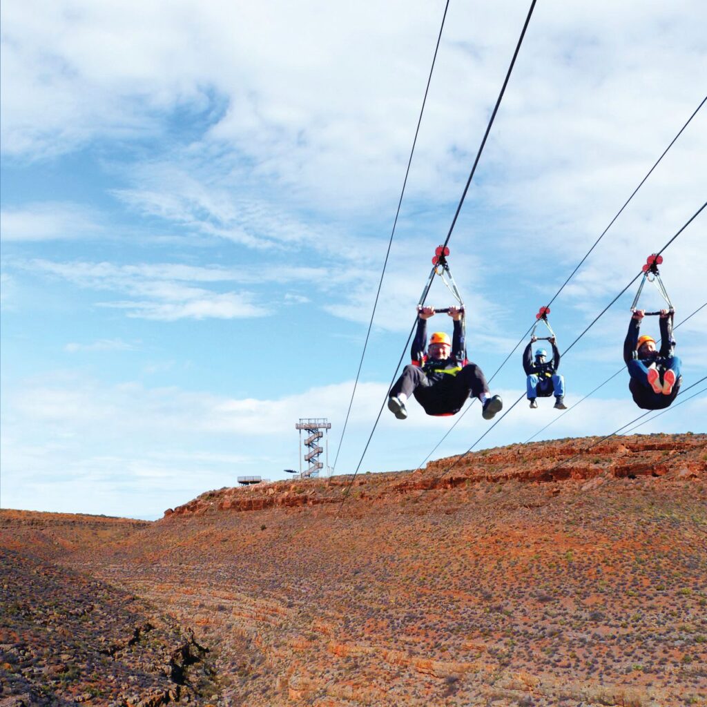 Three individuals, wearing helmets and harnesses, are suspended in the air while riding parallel ziplines. They appear to be in mid-flight over a canyon or gorge.