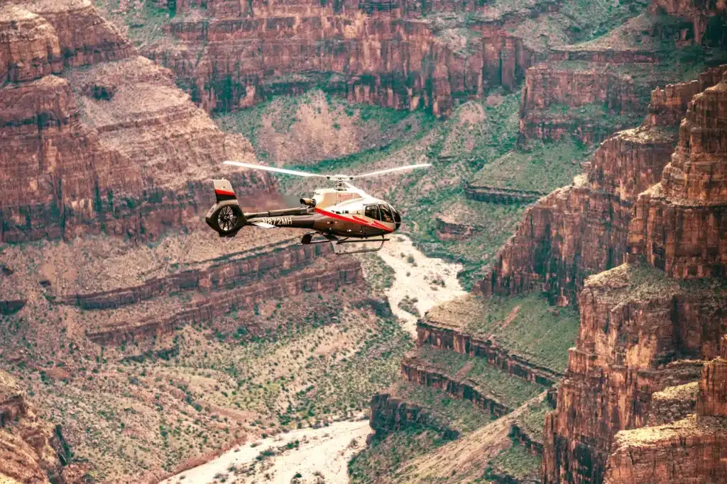 Red and white helicopter soaring above the rugged cliffs and winding river of the Grand Canyon, showcasing breathtaking aerial views of the canyon’s natural beauty.
