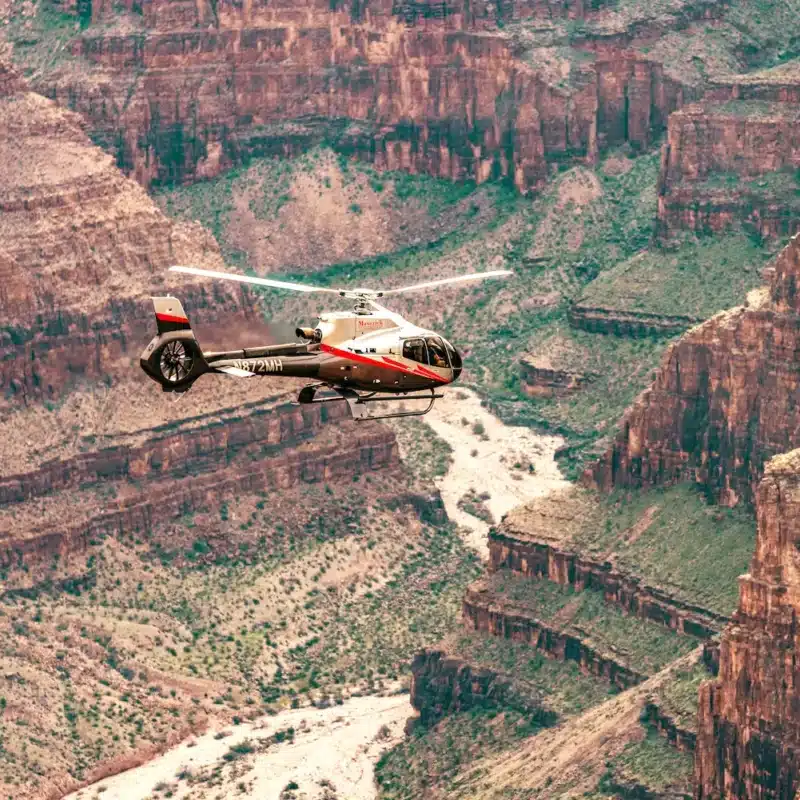 Red and white helicopter soaring above the rugged cliffs and winding river of the Grand Canyon, showcasing breathtaking aerial views of the canyon’s natural beauty.