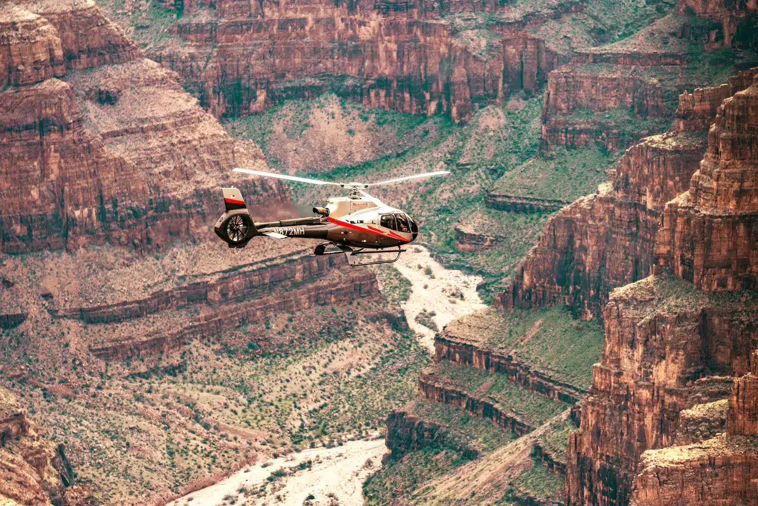 Red and white helicopter soaring above the rugged cliffs and winding river of the Grand Canyon, showcasing breathtaking aerial views of the canyon’s natural beauty.