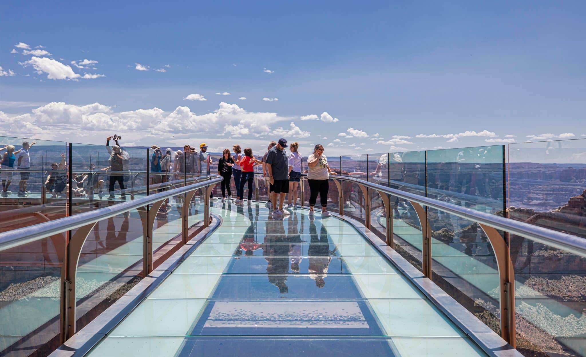 A group of tourists is walking on the U-shaped, glass-floored Skywalk over the edge of the Grand Canyon, which offers panoramic views of the distant canyon landscape under a bright blue, cloudy sky.