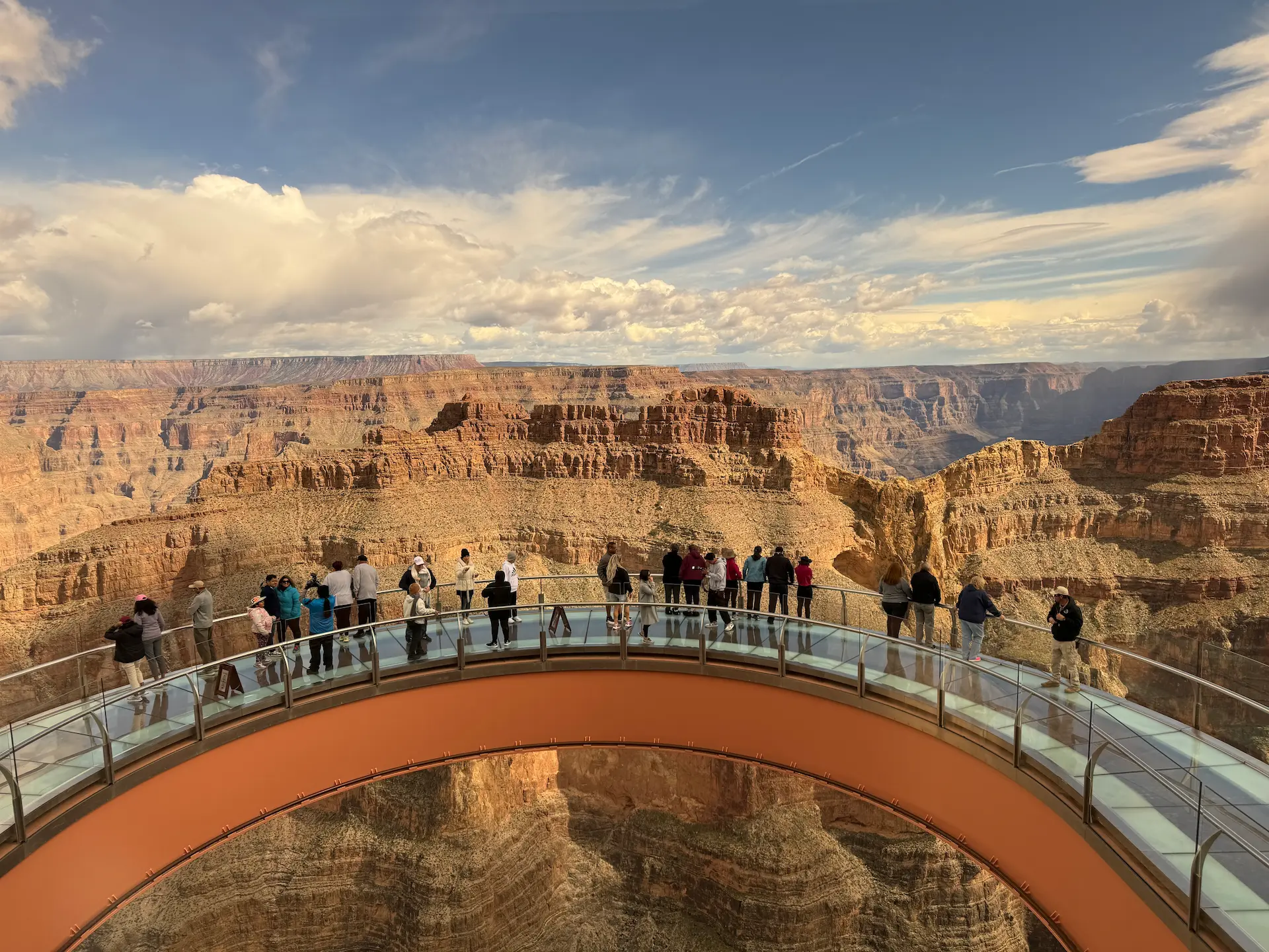 Tourists walking on the glass bridge of the Grand Canyon Skywalk with a panoramic view of the canyon rock formations and cloudy sky.