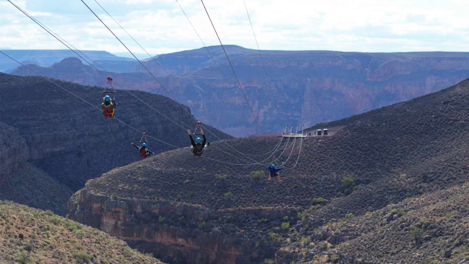 Zipline Feature Four people are seen zip-lining across a deep, V-shaped canyon gorge with steep, rocky walls, against a backdrop of distant, hazy blue mountains.