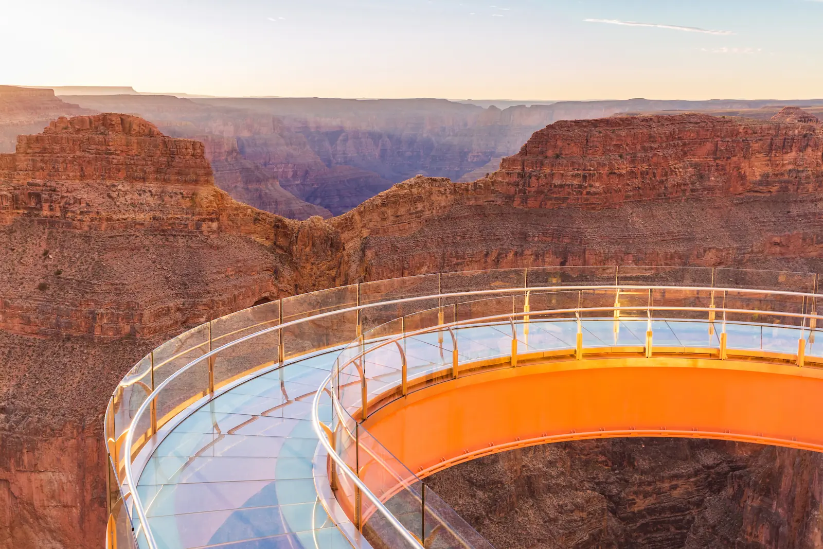 Curved glass-bottom Grand Canyon Skywalk reflecting warm sunset light, overlooking dramatic canyon cliffs and deep gorge in the background.
