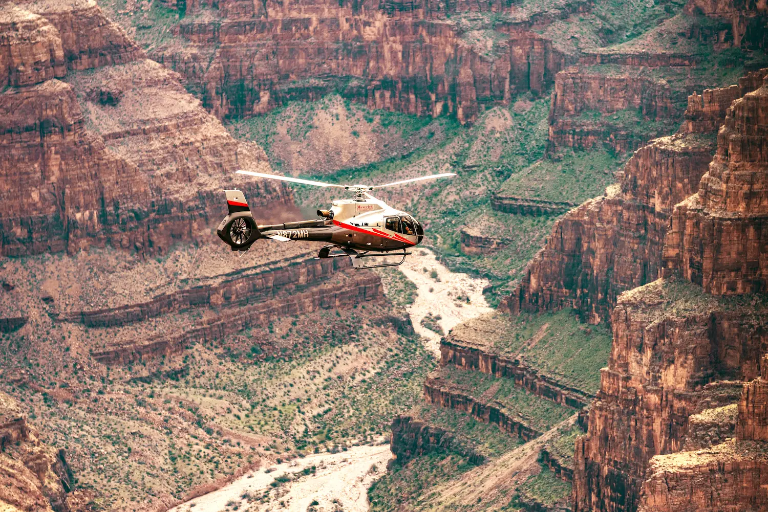 Red and white helicopter soaring above the rugged cliffs and winding river of the Grand Canyon, showcasing breathtaking aerial views of the canyon’s natural beauty.