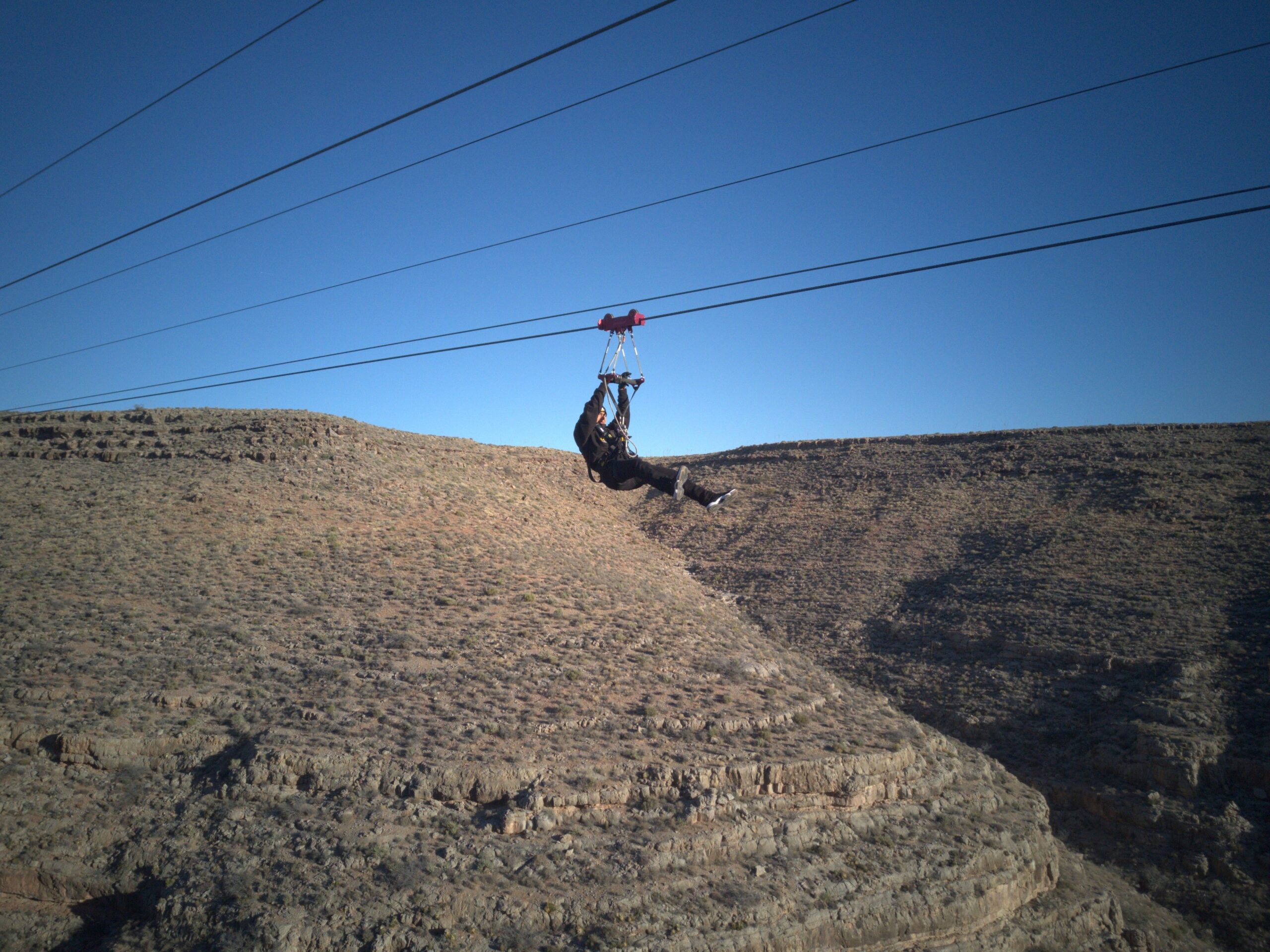 Zipline Feature A person in dark clothing and a helmet is seen mid-flight on a zipline, suspended over a rugged, rocky canyon beneath a clear blue sky.