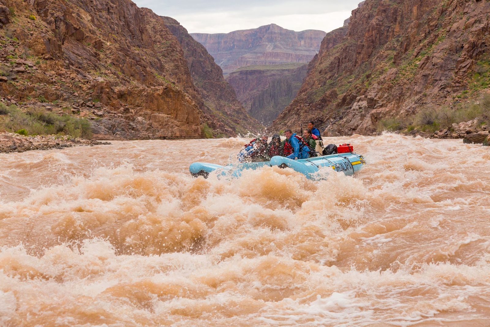 Phoenix to Grand Canyon Road-Trip Guide A raft carrying several people navigates turbulent, muddy brown rapids on a river in a deep canyon surrounded by towering, reddish-brown rock walls.