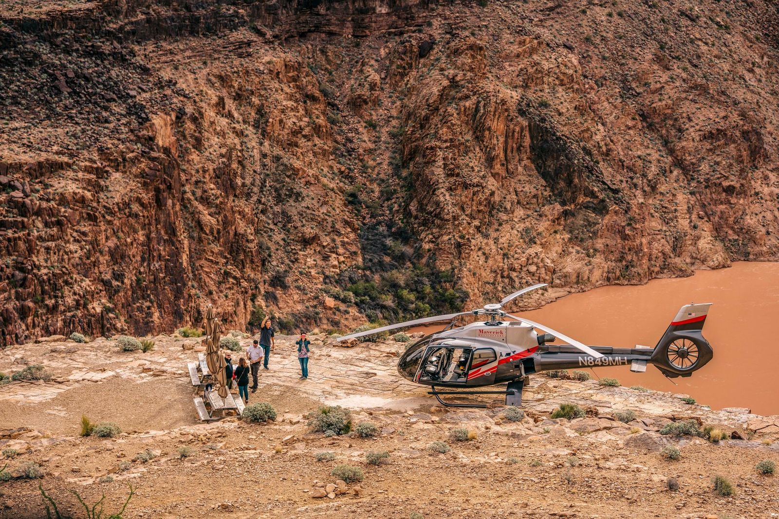 Phoenix to Grand Canyon Road-Trip Guide A gray and red Maverick helicopter is landed on a dusty, rocky ledge next to a muddy, orange-brown river, while a small group of tourists walks nearby against a backdrop of steep, reddish-brown canyon walls.