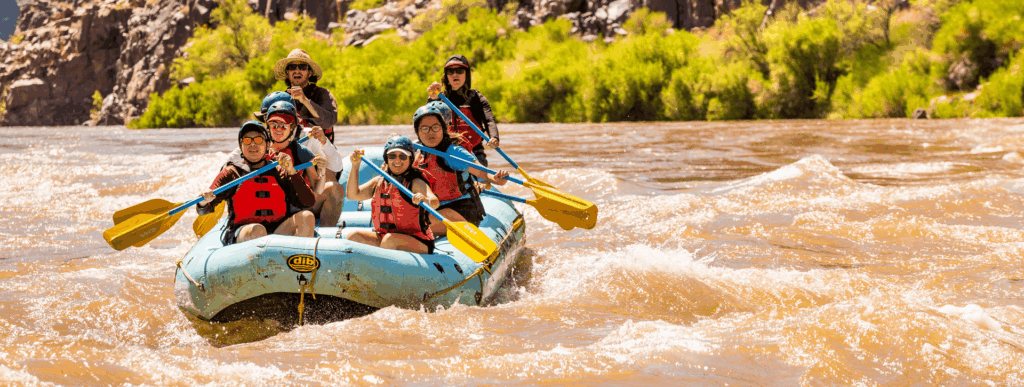 A group of people in a raft white water river rafting through rapids on a muddy river.