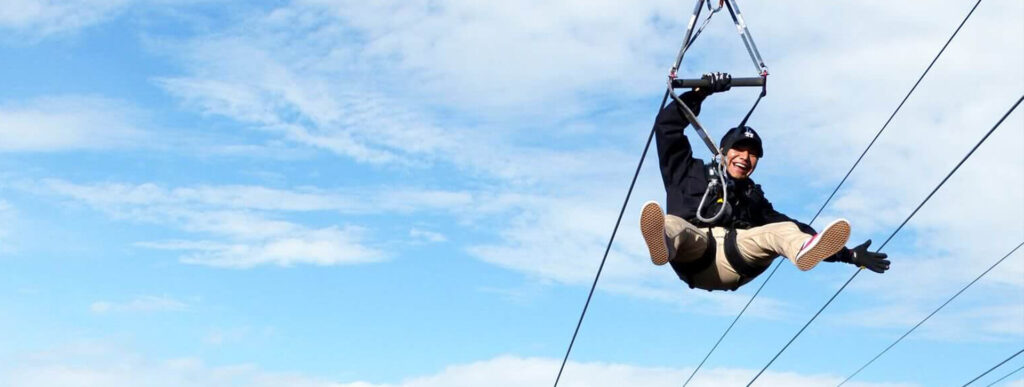 Dinge die zu tun sind A person smiles while zip-lining against a blue, cloudy sky.